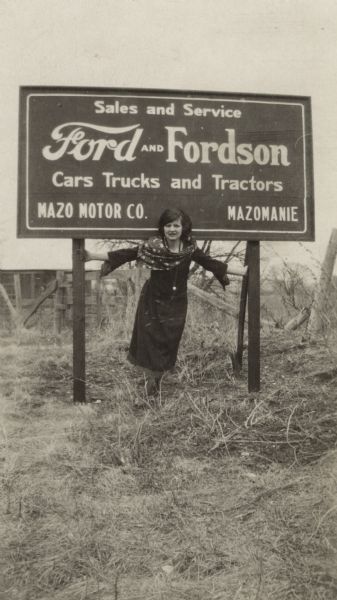 A woman in a dress and scarf poses beneath a Ford and Fordson sign. Farm buildings are in the background.