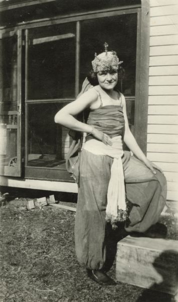 A woman in a costume gazes into the camera with one foot resting on a box, one hand on her hip, the other hand on her knee. Her costume consists of harem trousers, a sash around her waist, a camisole top and a hat that looks like a crown with a veil attached to the back. She is wearing stockings without shoes. A porch is behind her.