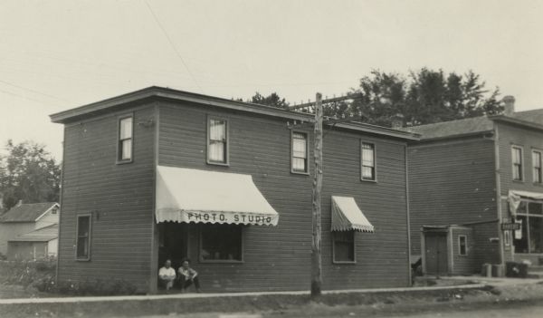 Matthew Witt's photo studio on Main Street. His business was there for 50 years, from 1913 to 1963. Two men are sitting on the stoop in the doorway. The building has two awnings. A bakery is next door on the right.
