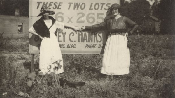 Two women pose in front of a sign advertising lots for sale. They are both dressed fashionably and their arms are stretched out with their fingers touching. One woman has a fur cape draped over her shoulders and the other is wearing a sheer blouse.