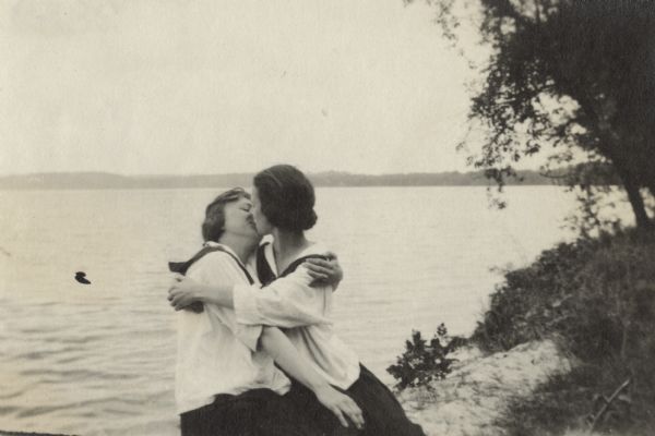 Two women embracing and kissing on the shore of a lake or river. They are both dressed in dark skirts and middy blouses with ties.