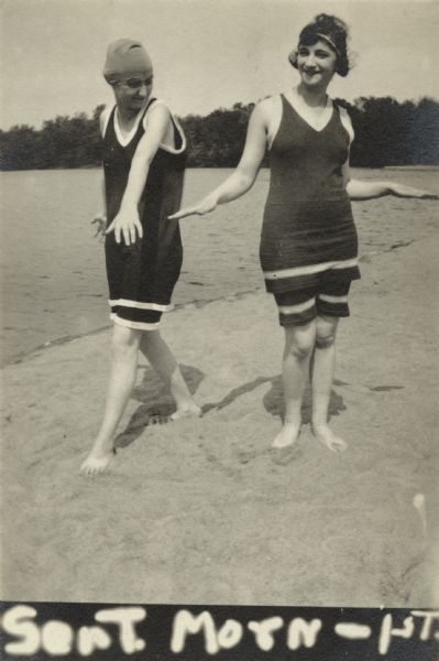 Two women strike a pose on the beach wearing bathing suits. One woman wears a bathing cap, the other a headband.