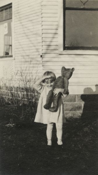 A young girl stands in a yard near the corner of a house with her Teddy Bear in one arm. She is shading her eyes from the sun and is looking down. She is wearing a dress, stockings and shoes.