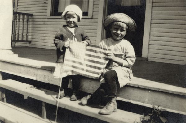 Two young children on the top step of a porch are holding an American Flag. One is standing and the other is sitting. They are wearing dresses, stockings, shoes and hats.