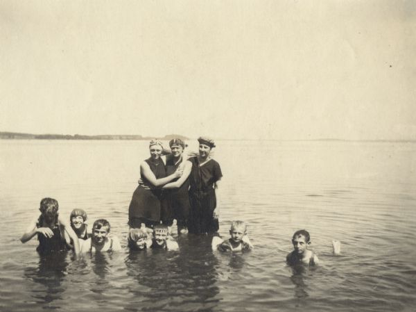 Seven young swimmers and a man crouch down in the water while three women wearing bathing suits and caps stand behind them. In the background is a shoreline.