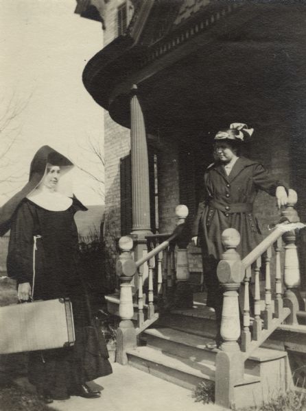 A nun stands on the sidewalk with a suitcase near a woman standing on the steps to a porch. The nun is wearing the traditional habit and headpiece and the woman is wearing a dress and hat.
