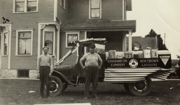 Roman Esser and Werner B. Esser stand in front of their truck. They are wearing work clothes. The truck appears to be decorated for a parade. A banner on the side reads, "Standard Oil Company (Indiana) and Red Crown Gasoline. Another sign reads, "Polarine Motor Oil, Made in Five Grades." Containers of products fill the truck bed. In the background is a house.