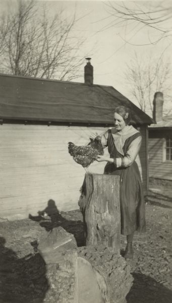 A woman stands in the yard petting and scratching a chicken perched on a stump. She is wearing a dress and has a barrette in her hair. In the background is a house and a shed.