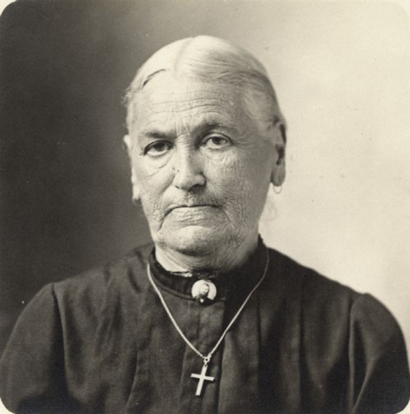 Quarter-length portrait of unidentified woman. She is wearing a dark dress, a pin with a man's image on it, and a crucifix on a chain.