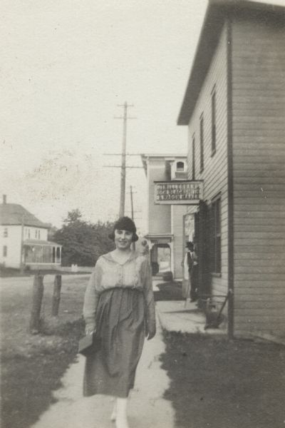 A smiling woman walks down the sidewalk in a small town. She is carrying a a box-shaped purse in her right hand, and is wearing a skirt and blouse. A house is across the street on the left, and storefronts are on the right. The sign on the business behind her reads (possibly), "JI HILLEBRAND, GEN BLACKSMITH, & WAGON MAKER." A man is leaning out of the door, watching her as a she walks by.