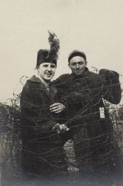 Portrait of a man and a woman posed together, wrapped in wire fencing. It must be a prank because they are both smiling. She is wearing a hat with a plume on the front, and a coat. He is wearing a jacket and cap and appears to have a pack on his back.