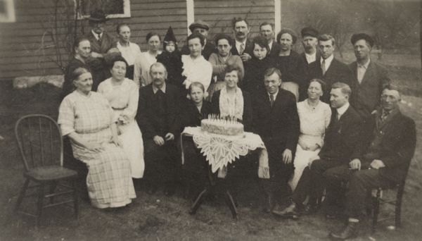 An outdoor group portrait of a birthday party. A cake with many candles and a lace tablecloth sits on a table in front. Most of the party are dressed up. Names are listed on back of photograph. Front row, Grandma Jacobs, Tina Braun, George Danz, unknown, Emma Straus, Fred Straus, Mazie Straus, Emil Straus and Ed Straus. Back row, Mrs. Barlow, John Barlow, Clarsie Barlow, Mandie Barlow, Mrs. Roy Sarbecher, Mrs. Walter Brookman, Walter Brookman, Mrs. Anna (Ed) Straus, Bill Danz, Tillie Braun (Mrs. Hugo Faust), unknown, Mary Grob (Mrs. George), Jonhn Braun, Karl Straus, George Grob.