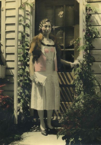 Ann Drucella Witt, the sister of Matt Witt, poses under a vine covered trellis that frames a door to a house. She is wearing a dress, jacket, gloves, and fur stole and is holding a purse. She worked with her brother in his photography business, and hand-tinted many photographs.