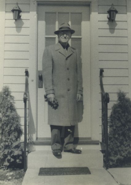 Matthew Witt, photographer, standing on the steps in front of the door to his second home. He is wearing an overcoat and hat, and is holding his gloves.