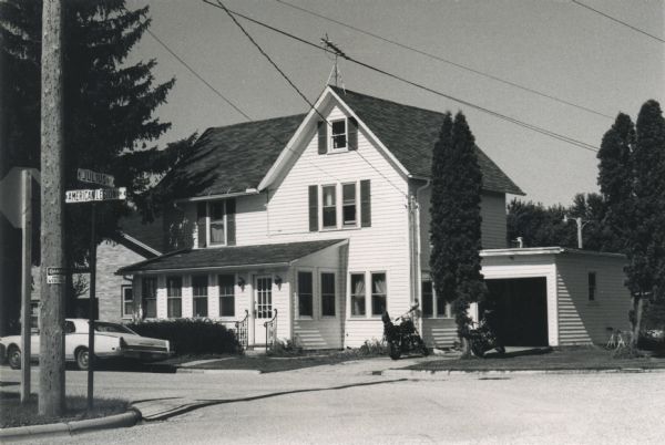 View from street of the second home of Matthew Witt, photographer, located on the corner of Julius Street and American Legion Drive. Two motorcycles are parked in the driveway and an automobile is parked at the curb.