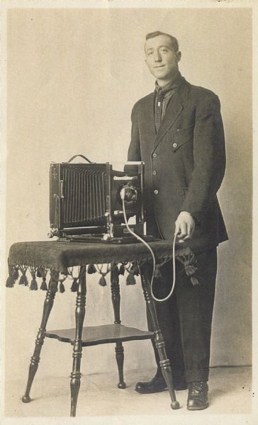 Photographer Matthew Witt poses with one of his cameras, holding the shutter release. It is placed on a table with a fringed tablecloth. He is wearing a suit.