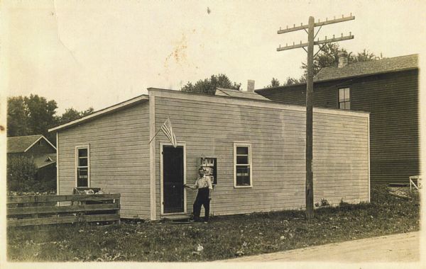 Hand-colored exterior of Matthew Witt's photography studio. He is standing at the door with his hand on the doorknob. Photographic prints are hanging in the window.