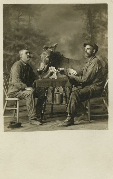 In a studio "gag" portrait, two men sit at a table playing cards in front of a painted backdrop. Both are showing their hands and smoking cigars. Each man has a glass of beer. A donkey stands behind the table. Both men are wearing work clothes.