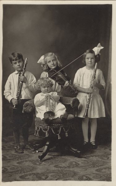 A full-length studio portrait of four children holding musical instruments, a violin, two clarinets and a trumpet. The youngest is seated in a chair. The three girls are wearing dresses.
