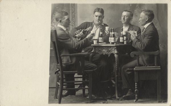 Photographic postcard of a full-length studio portrait in front of a painted backdrop of four men sitting at a table playing cards, smoking cigarettes and drinking alcohol.