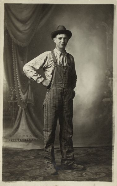 Full-length studio portrait of a man posing in front of a painted backdrop. He has one hand on his hip and is wearing striped overalls and a hat.