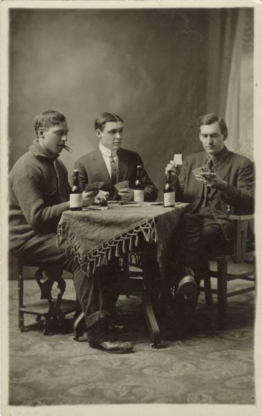 Studio portrait of three men, seated at a table, playing cards in front of a painted backdrop. Each man has a bottle of beer and one man smokes a cigar. Two men are wearing suits, the other on the left wears a sweater.