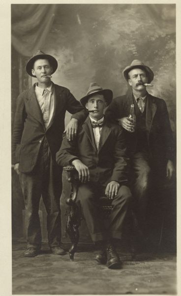 Full-length studio portrait of three men in front of a painted backdrop. Two men are standing on either side of a man in the middle who is sitting in a chair. They are all wearing suits, hats and smoking cigars. The man on the right holds a beer bottle.