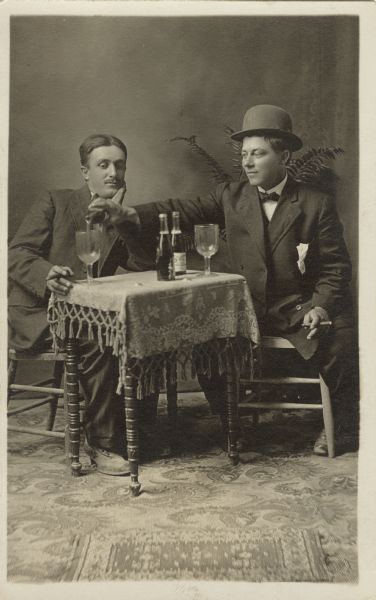 Studio portrait of two men, seated at a small table drinking. They are dressed in suits and each man is smoking a cigar. The man on the right is wearing a hat and is pouring a beer into the other man's glass. The man on the left has his chin in his hand and is watching his glass being filled. A fern is in the background.