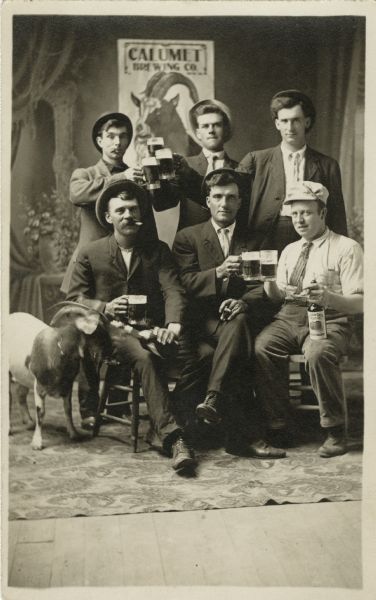 Full-length studio portrait of six men, three standing and three sitting. On the left is a goat. Attached to the painted backdrop, a poster for the Calumet Brewing Company, featuring a goat, can be seen behind the men. Most of the men are wearing suits, and all are wearings hats and holding beer mugs. Two groups are clinking their mugs in a toast. The seated man on the right is holding a bottle of Golden Gate Fruit Company's Apricot Brandy on his knee.