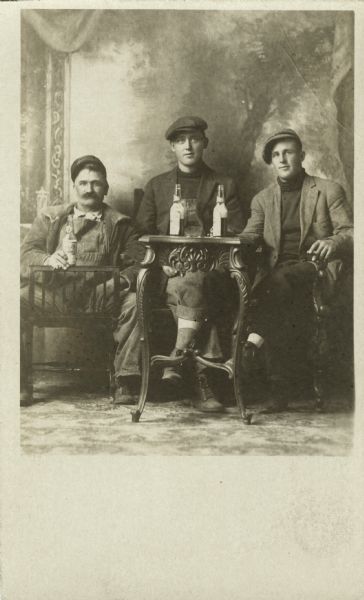 Studio portrait in front of a painted backdrop of three men, all seated in chairs around a table, drinking a beer from a bottle. The man on the right holds a cigar. They are dressed in casual or work clothes and hats.