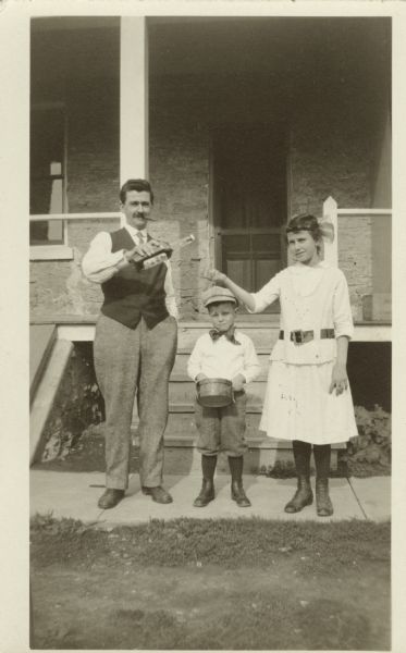 Group of three people standing outside on a sidewalk in front of a stone dwelling with a porch and steps behind them. A man holds a bottle, ready to pour into a glass held by a young lady. Between them is a boy holding a drum. He is dressed in knickers, stockings, shoes, shirt, bow tie and cap. She is wearing a dress with a belt, stockings, shoes and has a bow in her hair. The man is wearing trousers, shoes, shirt, vest and tie. He is smoking a cigar.