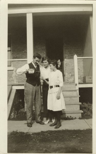 Group of three people standing outside on a sidewalk in front of a stone dwelling with a porch and steps behind them. A man holds a bottle, ready to pour into a glass held by a young lady. Watching between them is a woman. She is dressed in a skirt and blouse, stockings and shoes. The young lady is wearing a dress with a belt, stockings, shoes and has a bow in her hair. The man is wearing trousers, shoes, shirt, vest and tie. He is smoking a cigar.