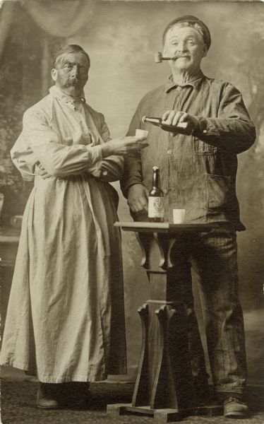 Full-length studio "gag" portrait of two men in front of a painted backdrop. The man on the right, James Gorman, is wearing work clothes, boots and hat and is smoking a corn cob pipe. He is holding a beer bottle (capped) as if to pour it into the cup of the other man. The man on the left (unidentified), appears to be wearing a long, very large, dress or apron. He has a bushy moustache and seems to have dirt on his face. A table stands in front with another beer bottle and cup.