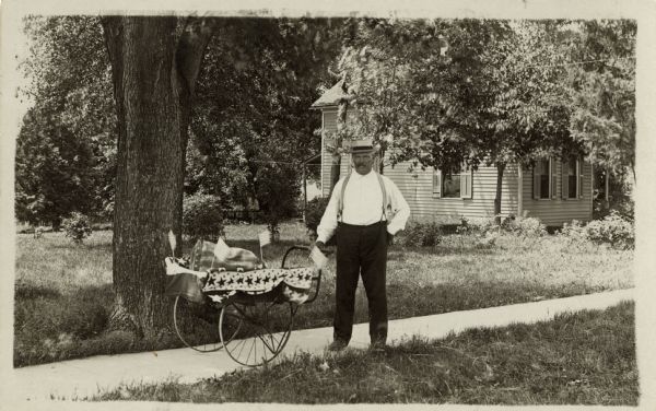 John Virnig, Sr., harness maker, grips the handle of a two wheeled hand cart as he poses on the sidewalk in a neighborhood. He is wearing a hat, shirt and pants with suspenders and has a moustache. A house and trees can be seen behind him. The cart is decorated with patriotic bunting and American flags. A striped bag of some kind is inside the cart.