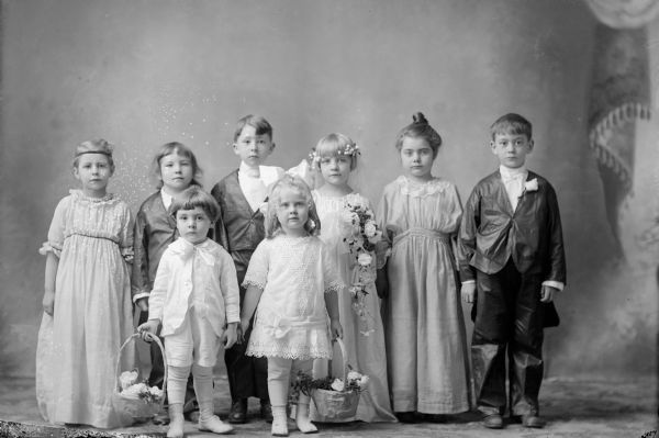 Studio portrait in front of a painted backdrop of a group of children. They are posed as a mock wedding party. The bride carries a bouquet and wears a circlet of flowers in her hair. The two bridesmaids wear dresses and the groom and groomsmen wear suits. In front, a girl and boy hold baskets of flowers.