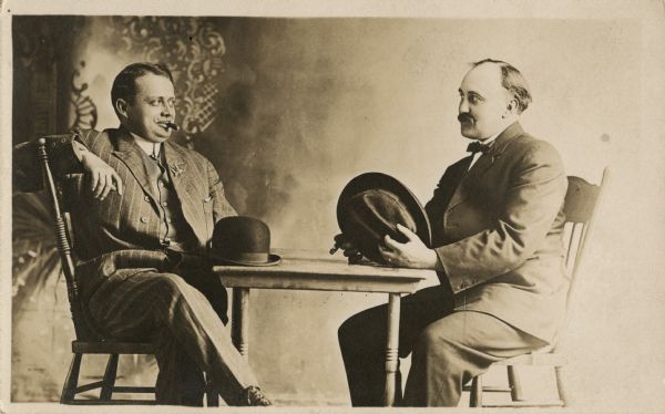 Studio portrait in front of a painted backdrop of two men seated at a table looking at each other. They are both wearing suits. The man on the left is smoking a cigar. His arm is resting on the back of his chair, his legs are crossed and his derby hat is on the table. The man on the right is holding his hat in his left hand and his cigar in his right.