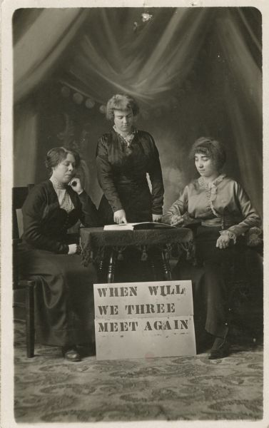 Studio portrait of three women surrounding a table with a book on it, in front of a painted backdrop. The middle woman is standing and pointing at the book, the two others are sitting in chairs on either side. All three are wearing dresses. A sign is propped against the table legs and it reads, "WHEN WILL WE THREE MEET AGAIN."