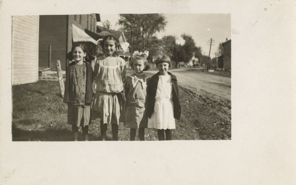 Four young girls pose outside of Witt's photography studio. They are wearing dresses. Two of the girls have jackets, and three of them have hair bows.