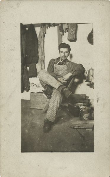 Man sitting on a box, leaning back against a wall, with his legs crossed. He is dressed in bib overalls and is smoking a pipe. He may be sitting in a kitchen as wood is stacked on the right and a coffeepot can be seen, possibly on a stove. Items are hanging on a rail overhead.