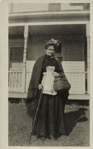 A woman poses on the lawn in front of a house with a porch. She is wearing a dress, apron, shawl, hat and shoes. In one hand she has a cane and in the other, a wicker basket with handles.