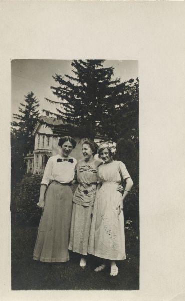 Three women pose outdoors. All are wearing dresses and one has a headband with a bow. Pine trees and a house can be seen in the background.