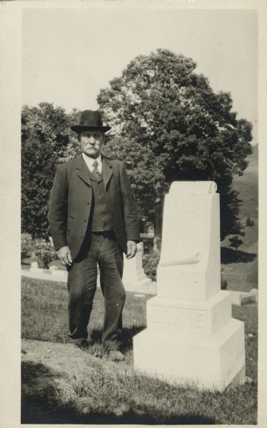 A man stands next to a headstone in a cemetery. He is wearing a suit and hat. The name on the headstone is "LOKKE."
