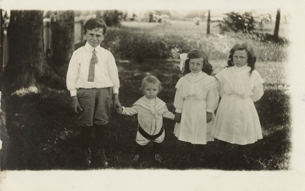 An older boy and two girls hold hands with a young boy. The girls are wearing dresses and the older boy is wearing knickers, stocking, shoes, shirt and tie. The young boy is wearing a sailor-style suit. A yard, fence and trees are in the background.