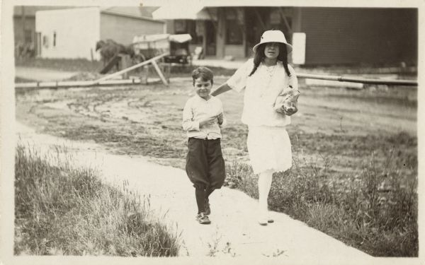 A boy and girl walk down a path. She has her hand on his shoulder, and is wearing a dress and hat carrying a sack. He is dressed in shirt, knickers, stockings and shoes. Commercial buildings and a horse-drawn wagon are in the background.