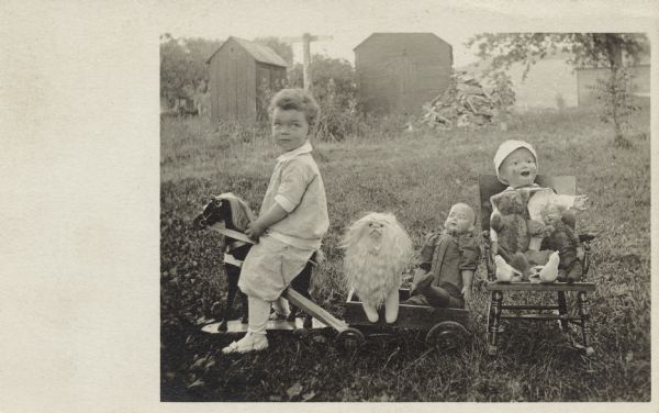 A child rides a hobby horse that is pulling a wagon filled with a toy lion and doll. A rocking chair holding a large doll, a small doll and teddy bear is placed behind the wagon. The boy is wearing knickers, shirt, stockings and shoes. Farm buildings are in the background.
