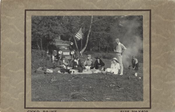 A group of men and women sit on the ground as they enjoy a picnic, eating and drinking. A dog is lying on the picnic blanket. An automobile with a flag mounted on it is parked behind them, and trees are in the background. A man on the far right appears to be tending a campfire.