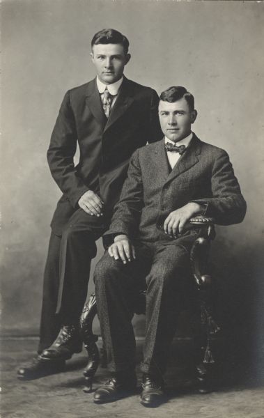 Full-length studio portrait of John Maly (seated) and another man (also with the last name Maly). One man is seated and the other is perched on the arm of the chair. They are both wearing suits, one with a tie and the other with a bow tie.