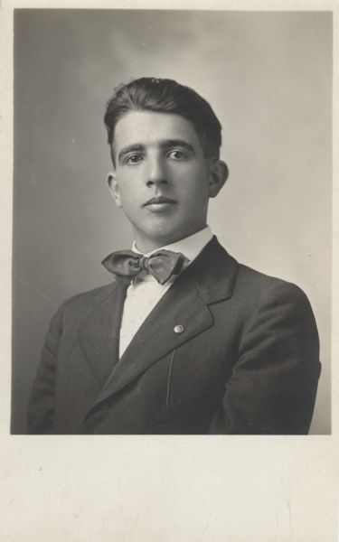 Quarter-length studio portrait of John Brunner. He is wearing a suit with a bow tie and a button with a chain in his lapel.