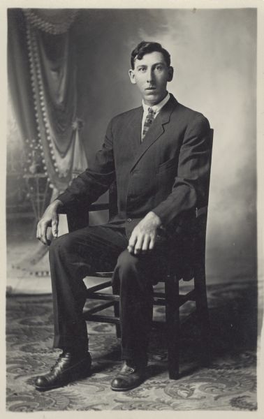 Full-length seated studio portrait of Chris Marking in front of a painted backdrop. He is wearing a suit.