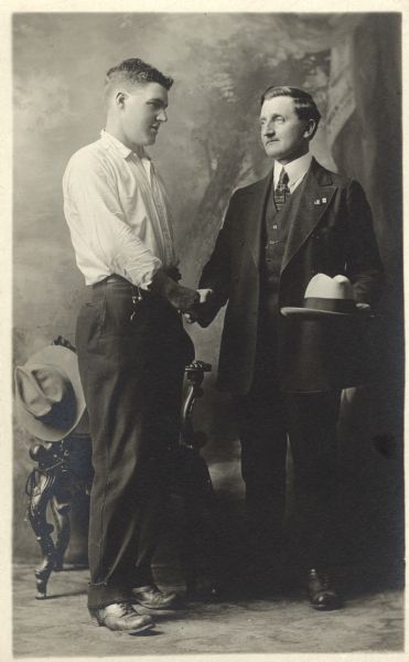 Full-length studio portrait in front of a painted backdrop of Ollie Bordson, wearing a suit, shaking hands with an unidentified man, who is wearing pants and a shirt. Ollie is holding his hat and and the other man's hat is hanging on the arm of a chair that is behind them.